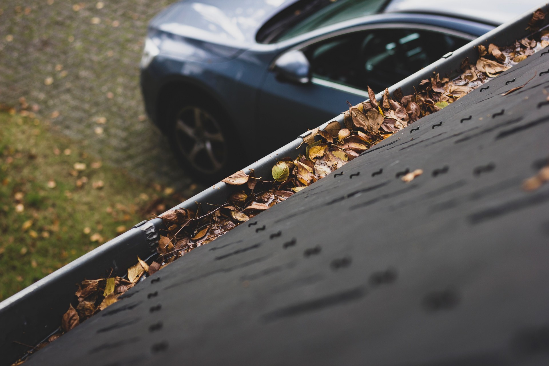 A portrait of a gutter and a slate roof of a house filled with fallen leaves due to autumn. The gutter needs to be cleaned during fall. In the background there is the driveway with a car on it.